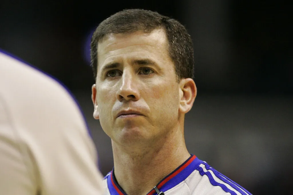 Close-up of a basketball referee wearing a gray uniform with blue and red trim, looking forward during a game