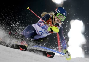 A skier in racing gear makes a sharp turn during a slalom run at night, kicking up snow as they lean low to the ground with bright stadium lights glowing in the background