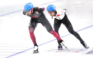 Two short-track speed skaters race side by side on the ice, one wearing a Canadian uniform and the other representing Japan, both leaning forward in a fast, aerodynamic stance
