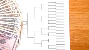 Blank tournament bracket on white paper next to a stack of U.S. dollar bills on a wooden table, symbolizing betting or prize pools.