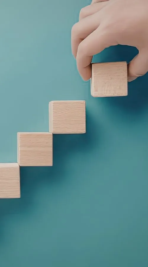 Hand placing a wooden block above other blocks arranged like steps against a teal background.