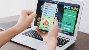 Person placing a sports bet on a smartphone beside a laptop displaying an online sportsbook.