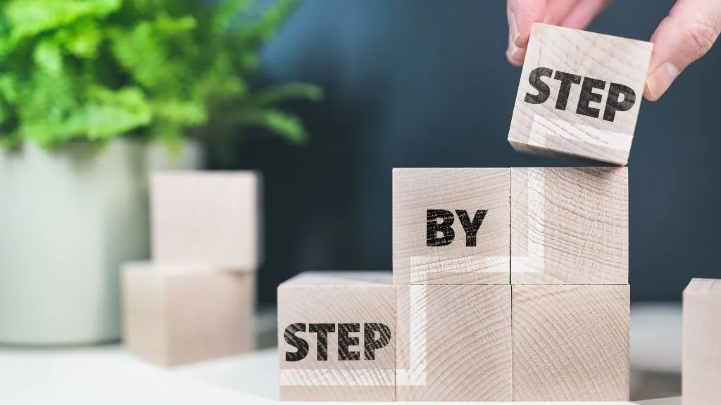 Hand stacking wooden blocks labeled “STEP BY STEP,” symbolizing a step-by-step process or guide
