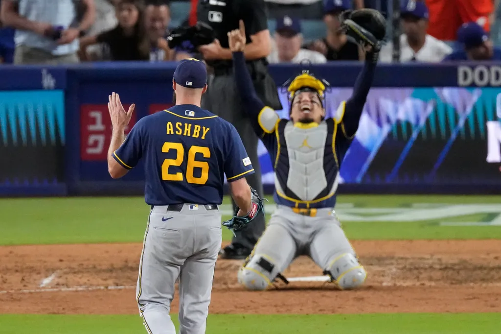 Milwaukee Brewers pitcher wearing “Ashby” and number 26 approaches a catcher celebrating on his knees with both arms raised.